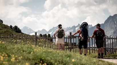 Drei Wanderer mit Rucksäcken blicken auf Bergpanorama hinter Holzzaun