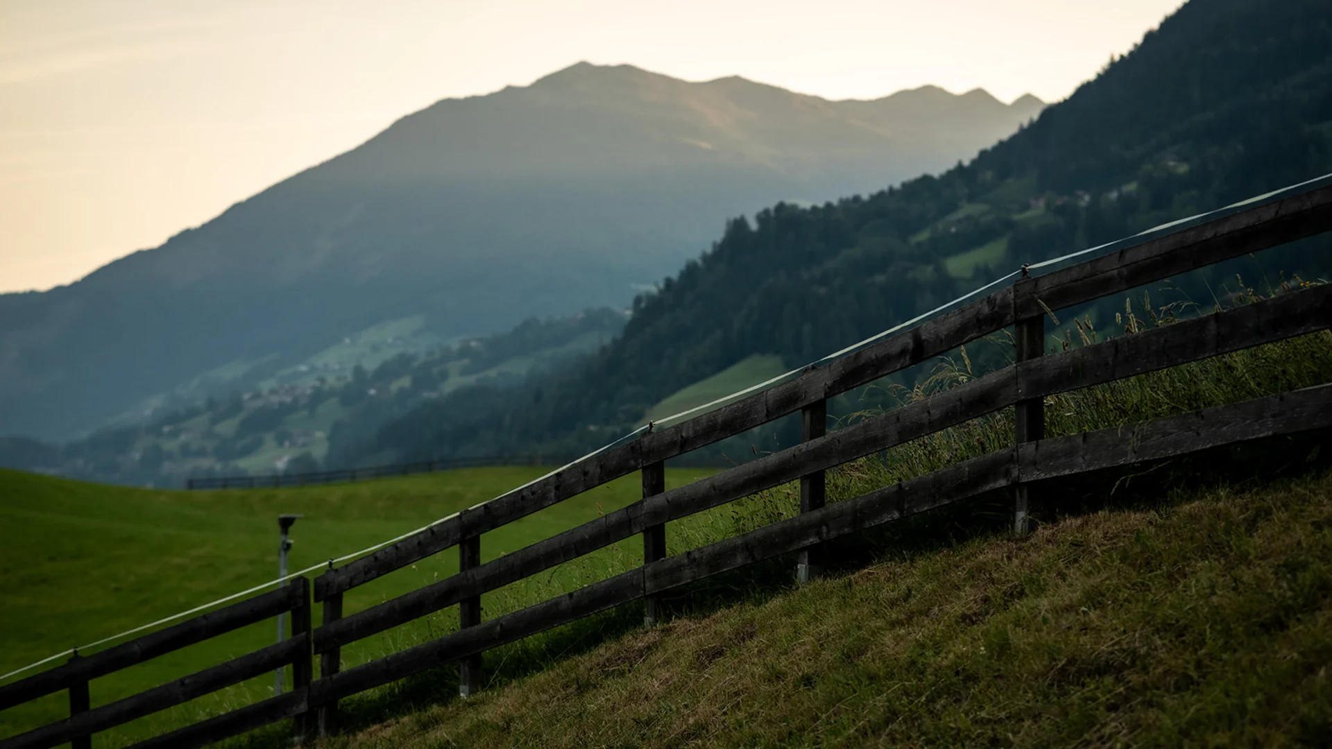 Holzzaun auf grasbewachsenem Hügel mit Bergen im Hintergrund bei Sonnenuntergang