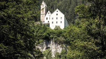 Weißes Schloss auf einem bewaldeten Felsen mit Berg im Hintergrund